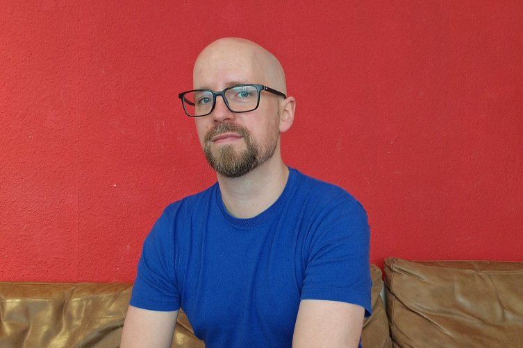 Robin Schmerer mit Brille und blauem T-Shirt auf einer Ledercouch vor einer roten Wand